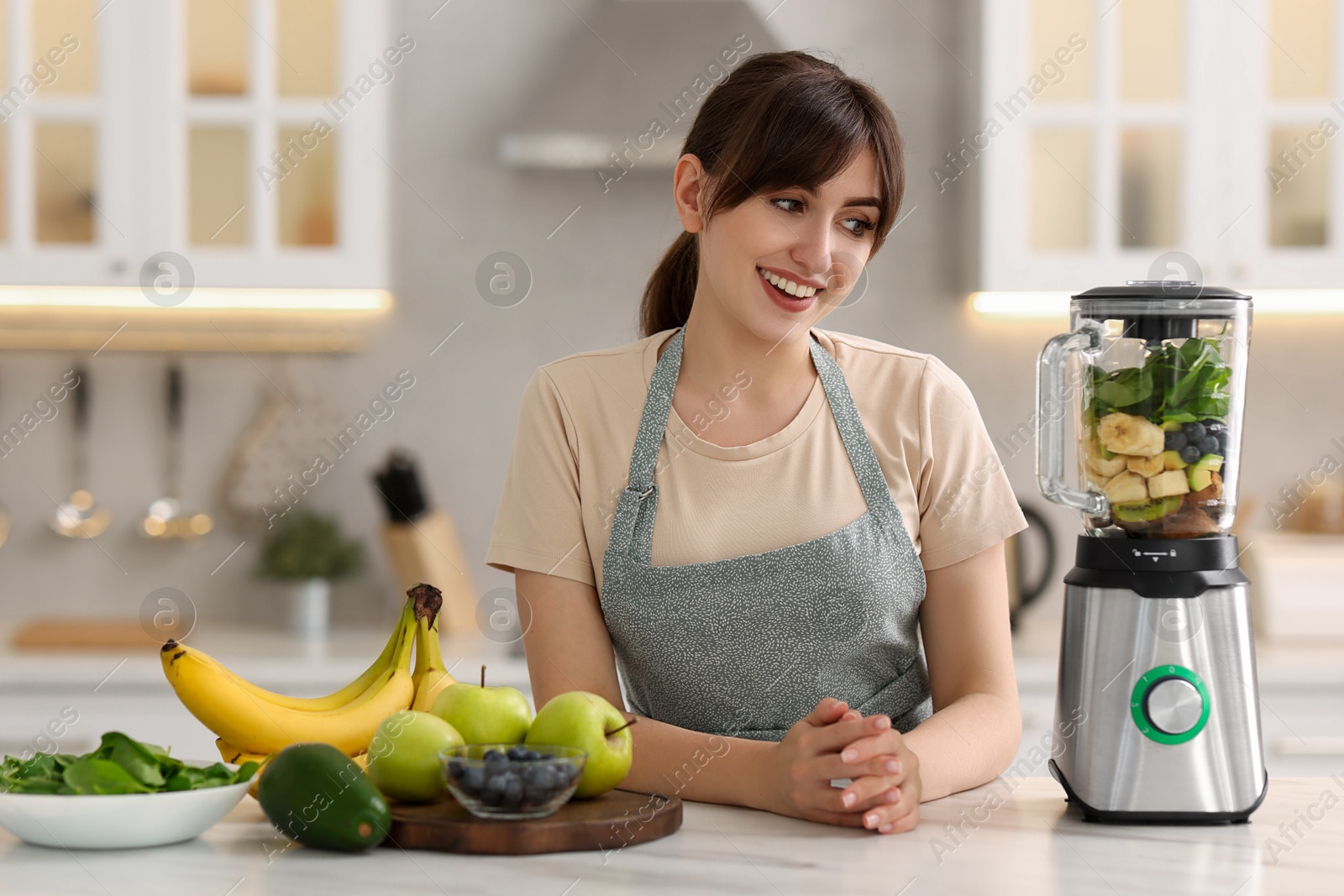 Photo of Young woman making delicious smoothie with blender at white marble table in kitchen