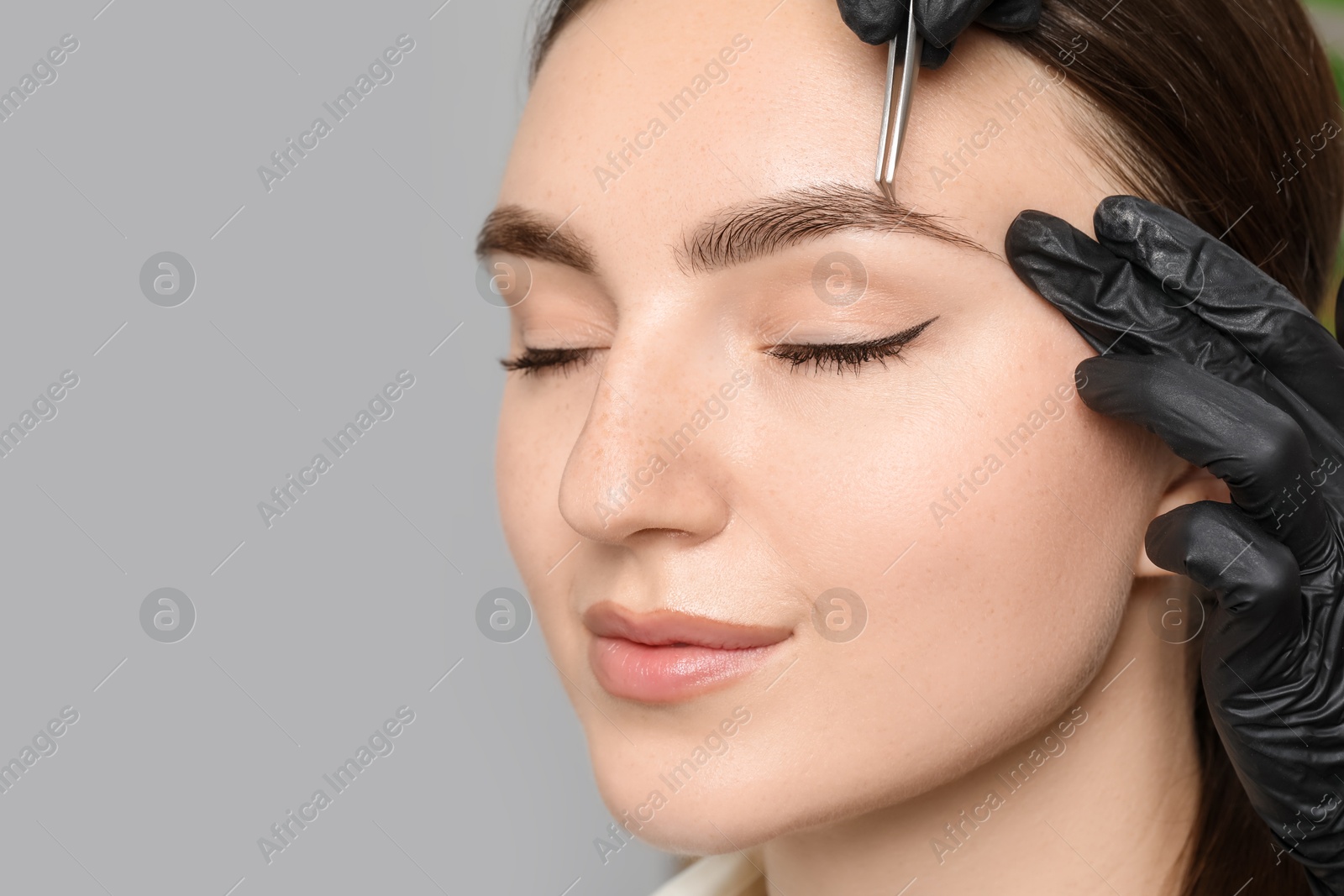 Photo of Beautician plucking young woman's eyebrow on light background, closeup and space for text