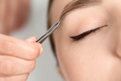 Photo of Beautician plucking young woman's eyebrow on light background, closeup