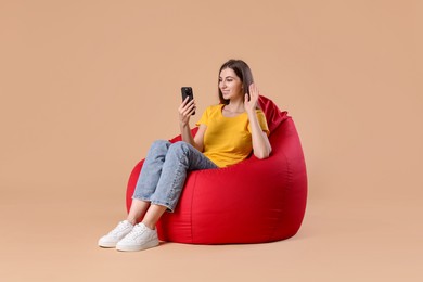 Photo of Smiling woman with smartphone having online meeting while sitting on red bean bag chair against beige background
