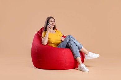 Photo of Smiling woman talking on smartphone while sitting on red bean bag chair against beige background