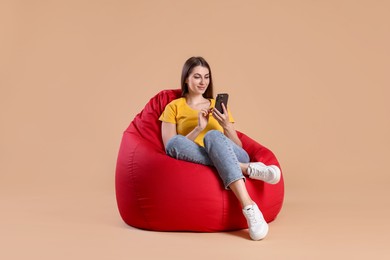 Photo of Beautiful young woman using smartphone on red bean bag chair against beige background