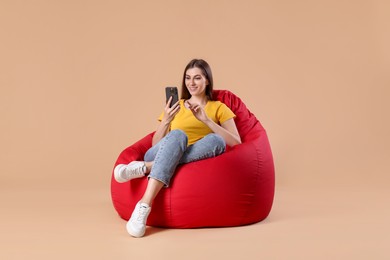 Photo of Smiling woman using smartphone on red bean bag chair against beige background