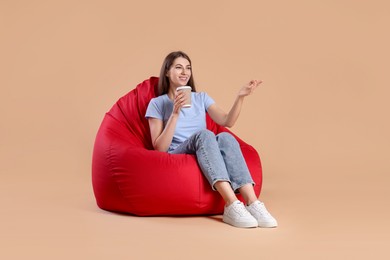 Photo of Smiling woman with paper cup of drink sitting on red bean bag chair against beige background