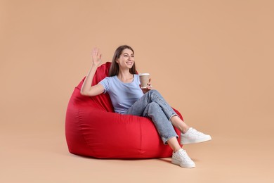Photo of Smiling woman with paper cup of drink sitting on red bean bag chair against beige background