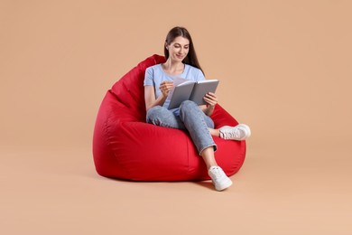 Photo of Beautiful young woman reading book on red bean bag chair against beige background