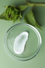 Petri dish with cosmetic product and sunflower on green background, flat lay