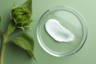 Photo of Petri dish with cosmetic product and sunflower on green background, flat lay