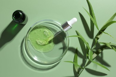 Photo of Petri dish with cosmetic product, bottle, pipette and leaves on green background, flat lay