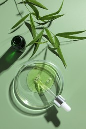 Petri dish with cosmetic product, bottle, pipette and leaves on green background, flat lay