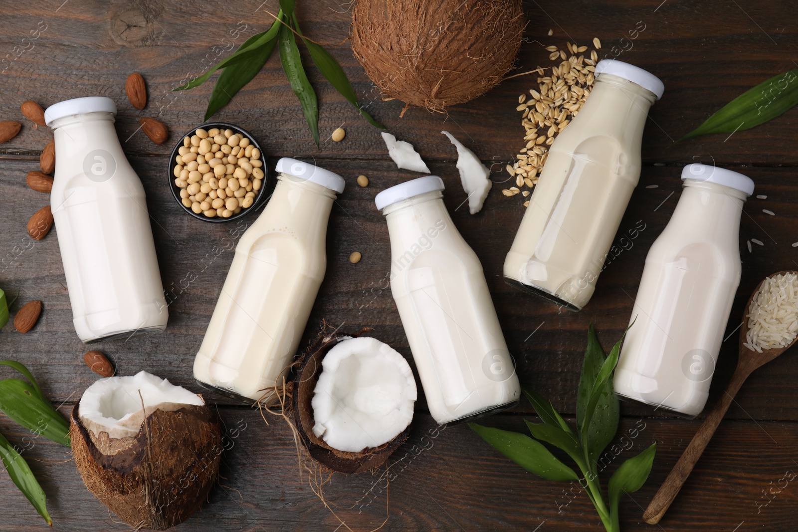 Photo of Different types of vegan milk in bottles with ingredients and green leaves on wooden table, flat lay