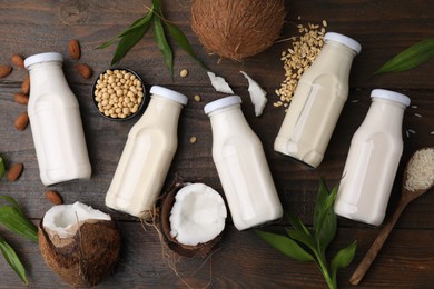 Different types of vegan milk in bottles with ingredients and green leaves on wooden table, flat lay