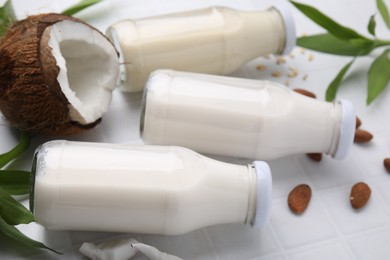 Different types of vegan milk in bottles and ingredients on white tiled table, closeup