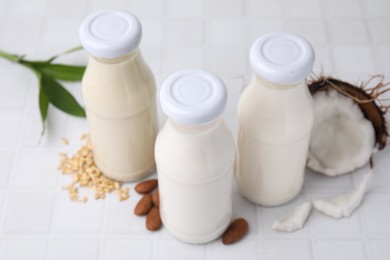 Different types of vegan milk in bottles and ingredients on white tiled table, closeup