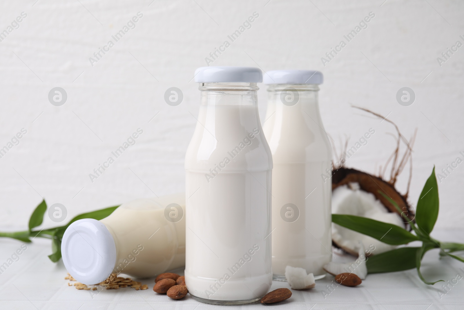 Photo of Different types of vegan milk in bottles with ingredients and green leaves on white tiled table