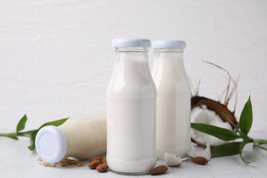 Different types of vegan milk in bottles with ingredients and green leaves on white tiled table