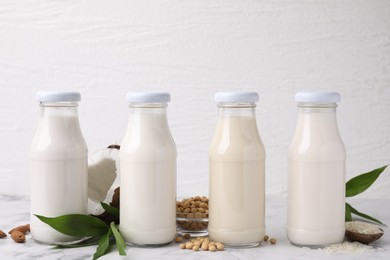 Different types of vegan milk in bottles with ingredients and green leaves on white marble table