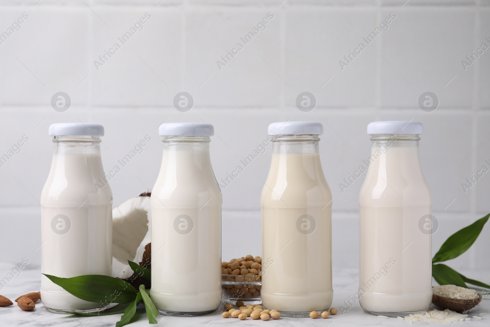 Photo of Different types of vegan milk in bottles with ingredients and green leaves on white marble table