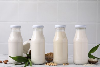 Different types of vegan milk in bottles with ingredients and green leaves on white marble table