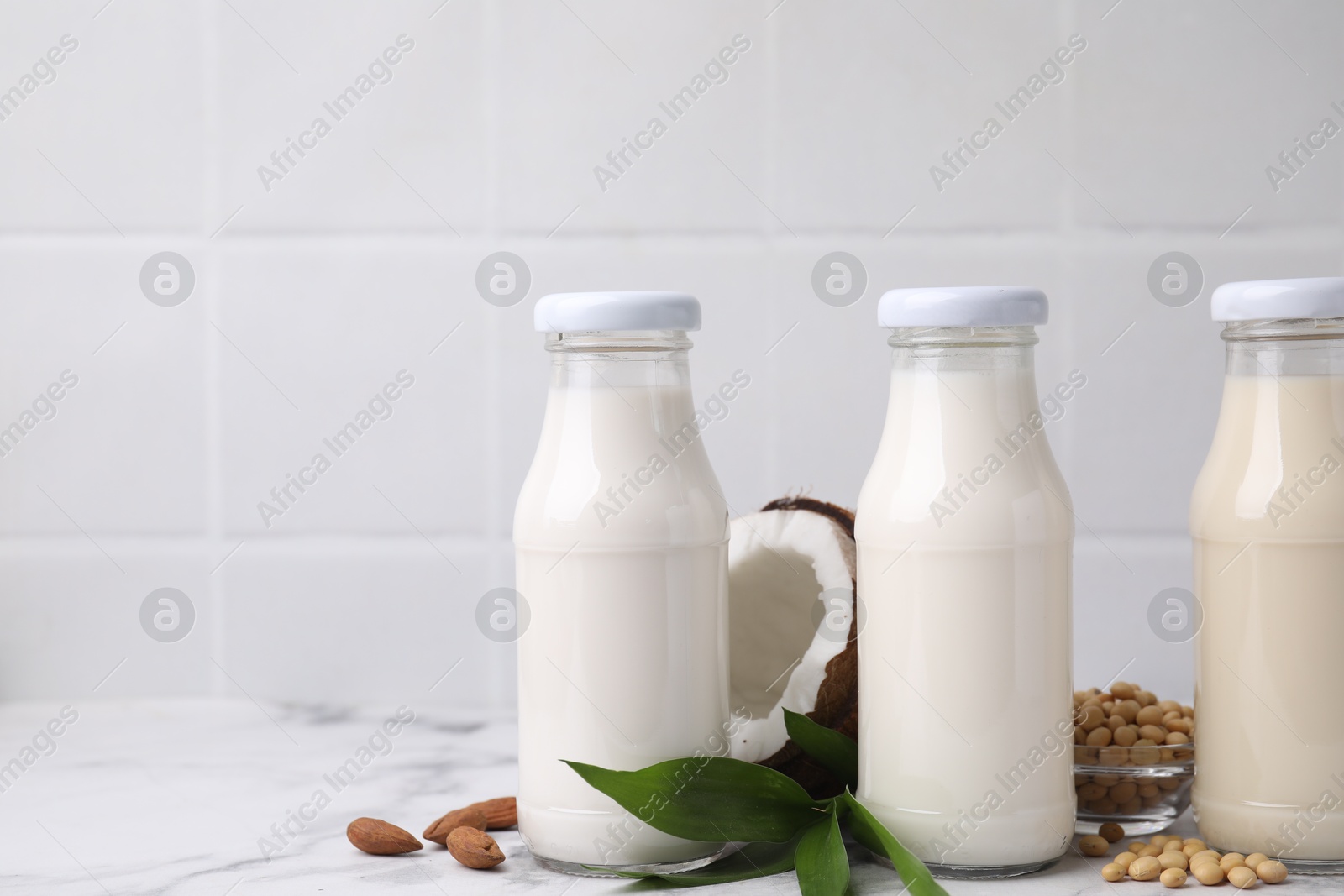 Photo of Different types of vegan milk in bottles with ingredients and green leaves on white marble table, space for text