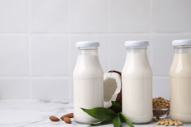 Different types of vegan milk in bottles with ingredients and green leaves on white marble table, space for text