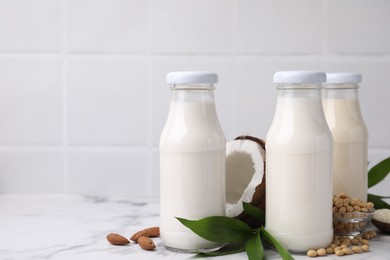 Different types of vegan milk in bottles with ingredients and green leaves on white marble table, space for text