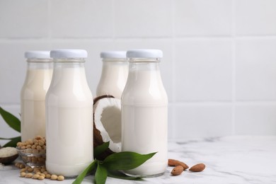 Photo of Different types of vegan milk in bottles with ingredients and green leaves on white marble table, space for text