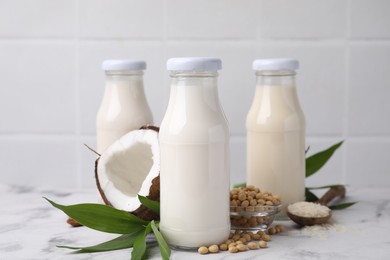 Photo of Different types of vegan milk in bottles with ingredients and green leaves on white marble table