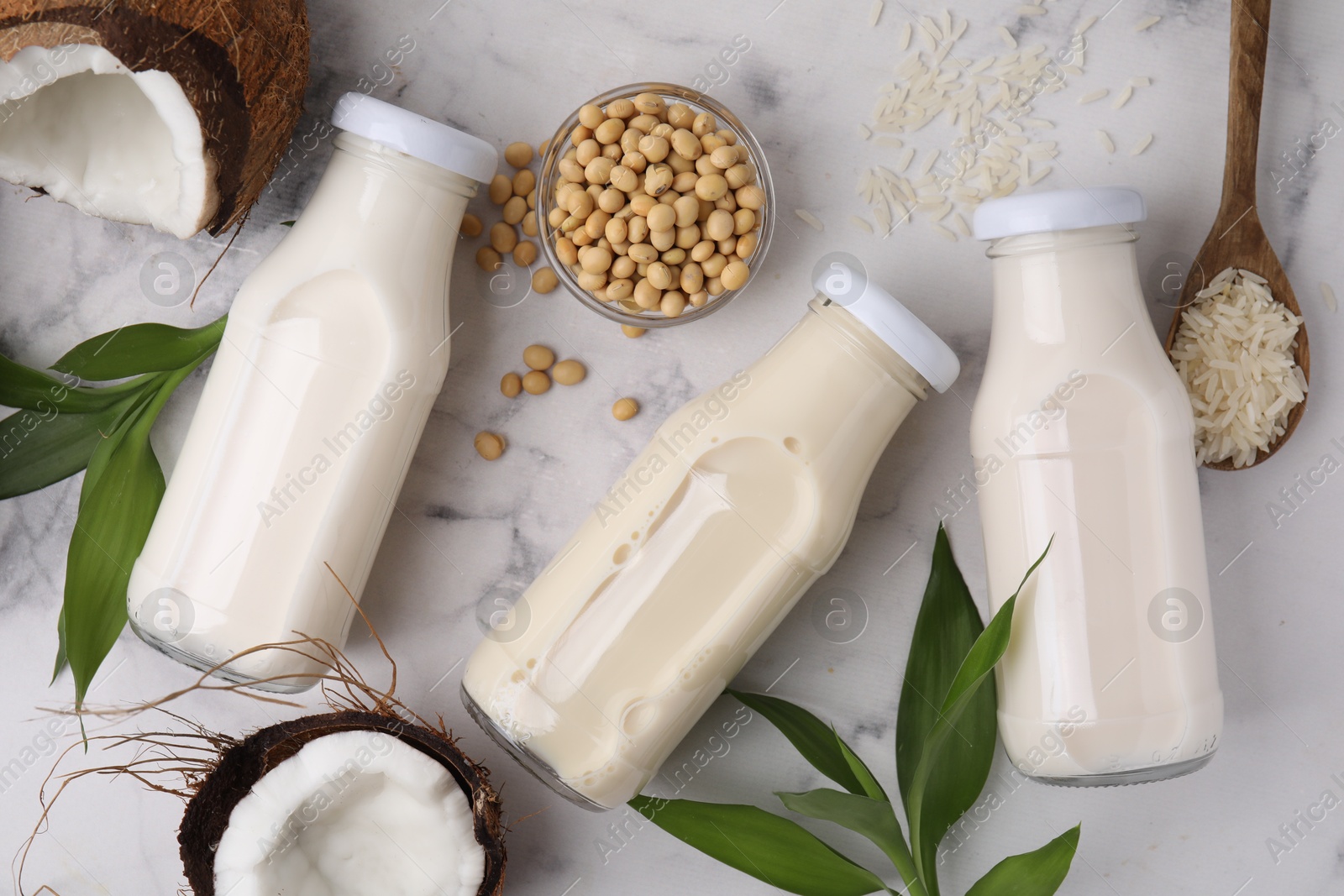 Photo of Different types of vegan milk in bottles with ingredients and green leaves on white marble table, flat lay