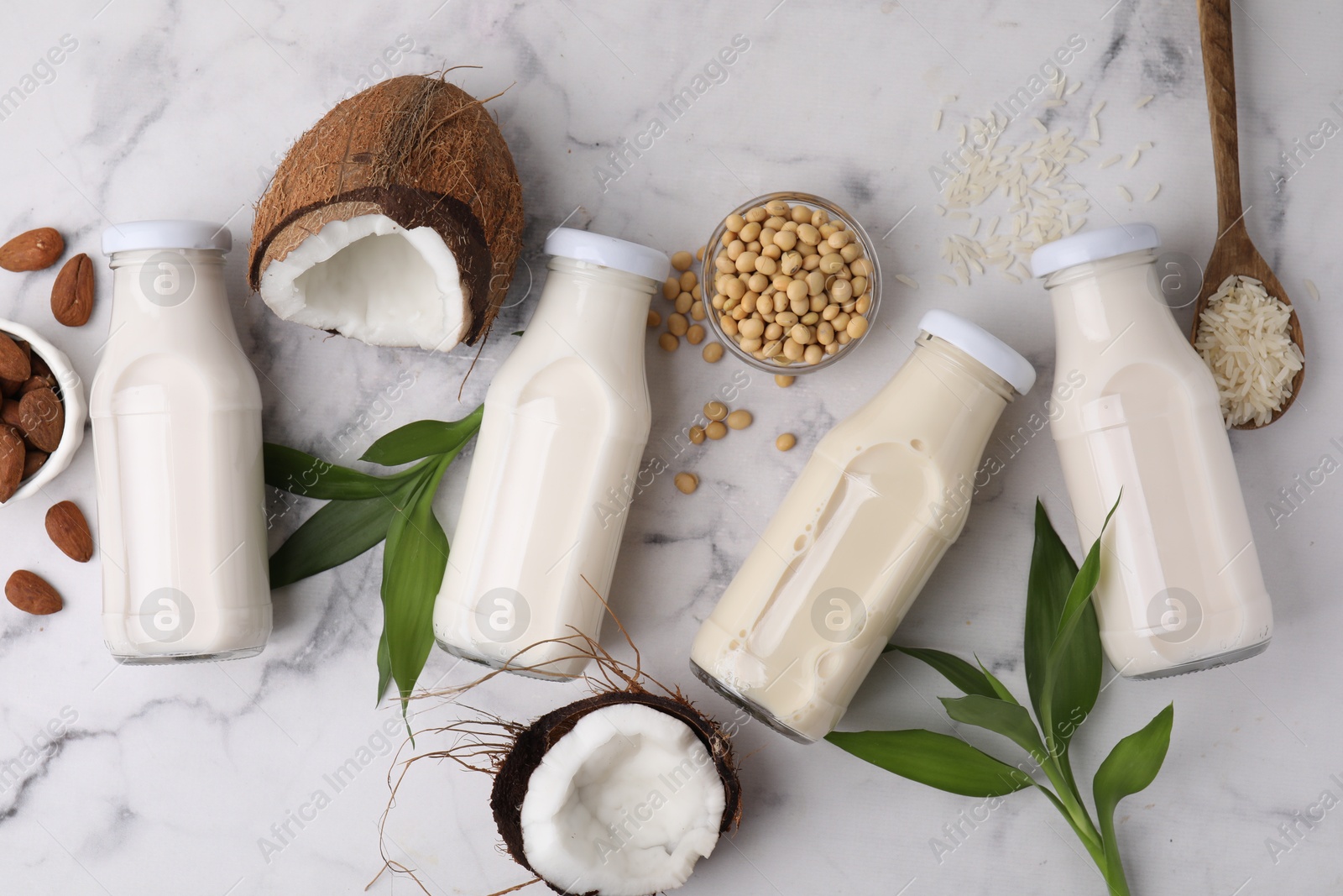 Photo of Different types of vegan milk in bottles with ingredients and green leaves on white marble table, flat lay