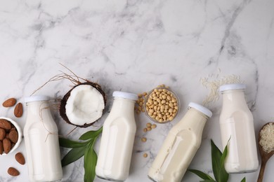 Different types of vegan milk in bottles with ingredients and leaves on white marble table, flat lay. Space for text