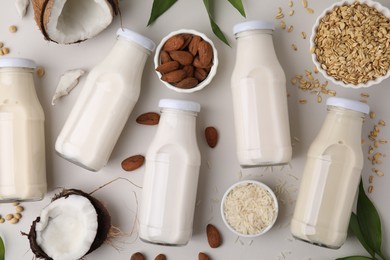 Different types of vegan milk in bottles with ingredients and leaves on light grey table, flat lay