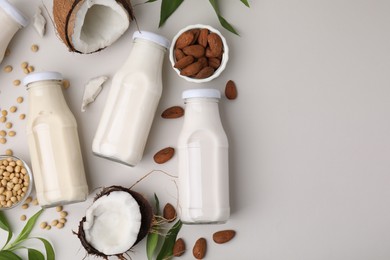 Different types of vegan milk in bottles with ingredients and leaves on light grey table, flat lay. Space for text