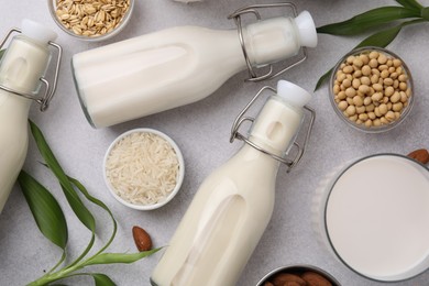 Different types of vegan milk with ingredients and leaves on light grey table, flat lay