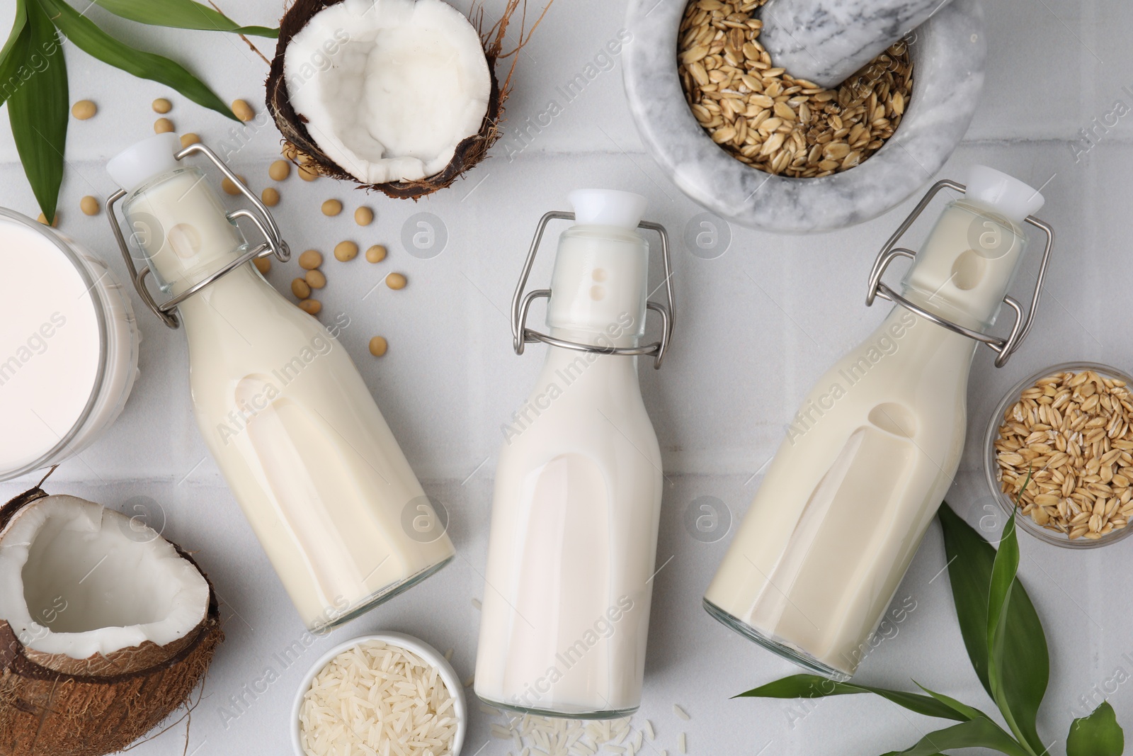 Photo of Different types of vegan milk in bottles with ingredients and green leaves on white tiled table, flat lay