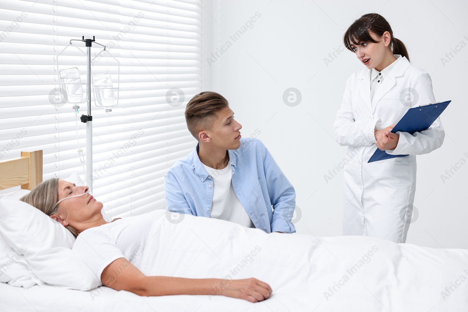 Photo of Coma patient. Sad young man near his unconscious mother talking with doctor in hospital