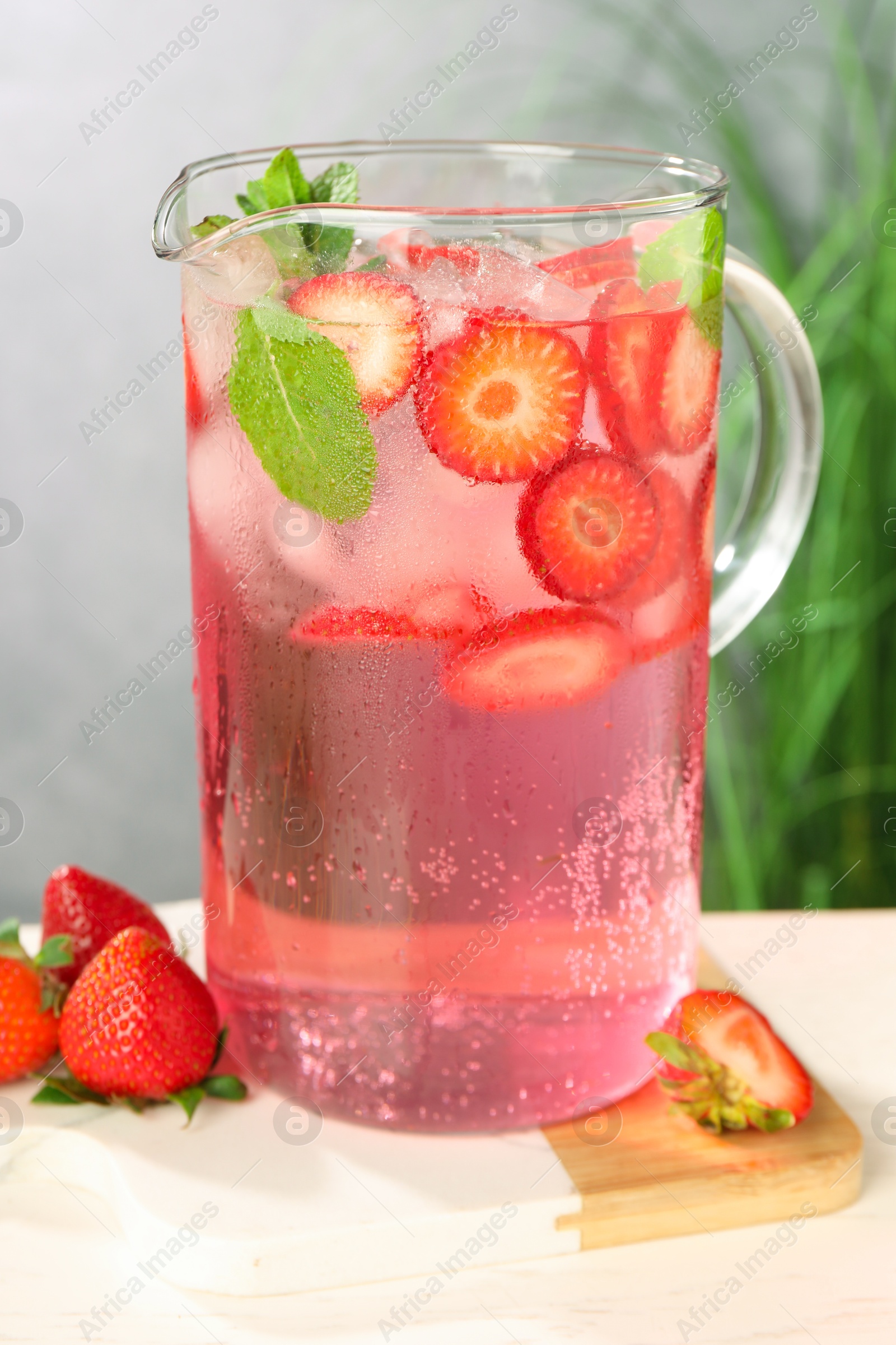 Photo of Freshly made strawberry lemonade with mint in jug and glass on white wooden table against grey background