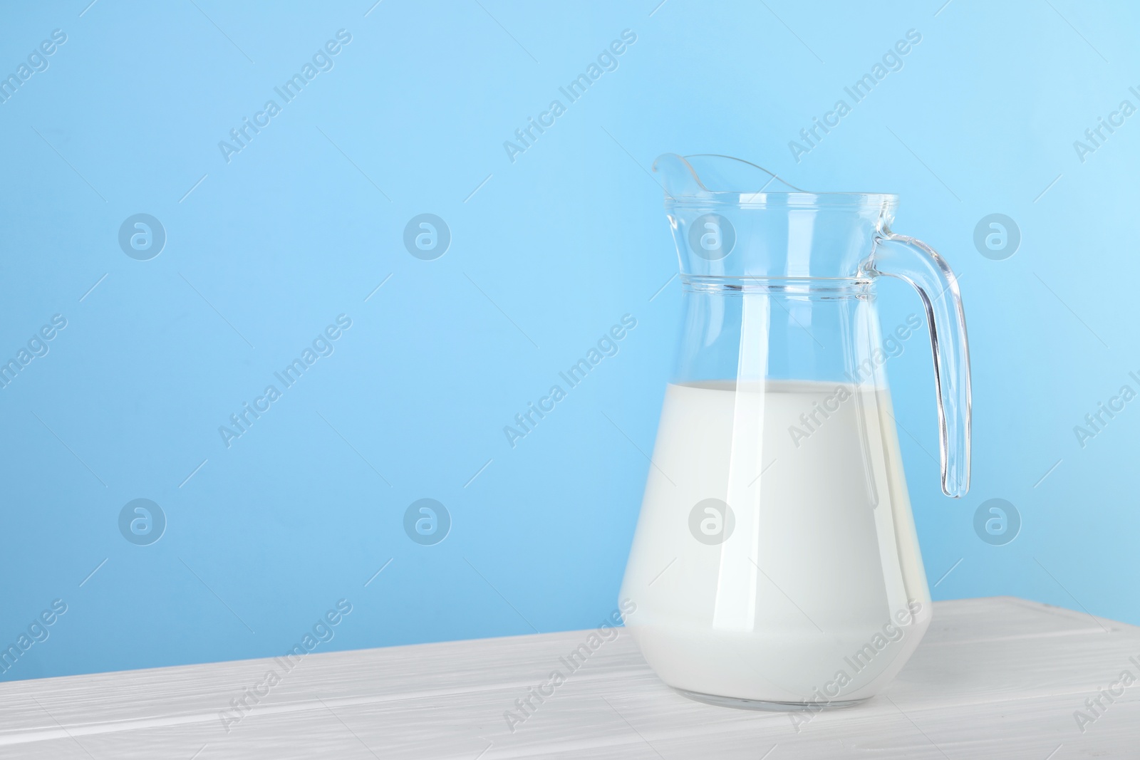 Photo of Jug of fresh milk on white wooden table against light blue background, space for text