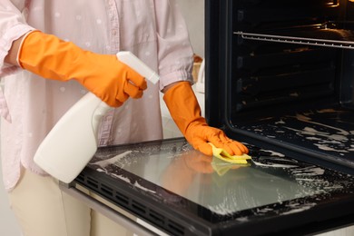 Photo of Woman cleaning oven door with rag and detergent in kitchen, closeup