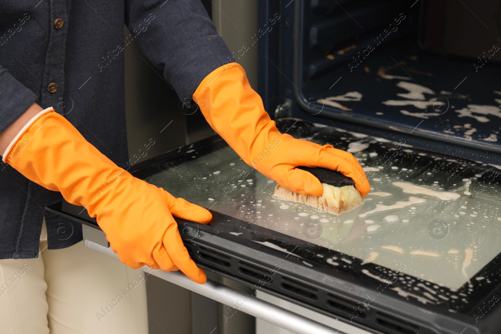 Photo of Woman cleaning oven door with sponge in kitchen, closeup