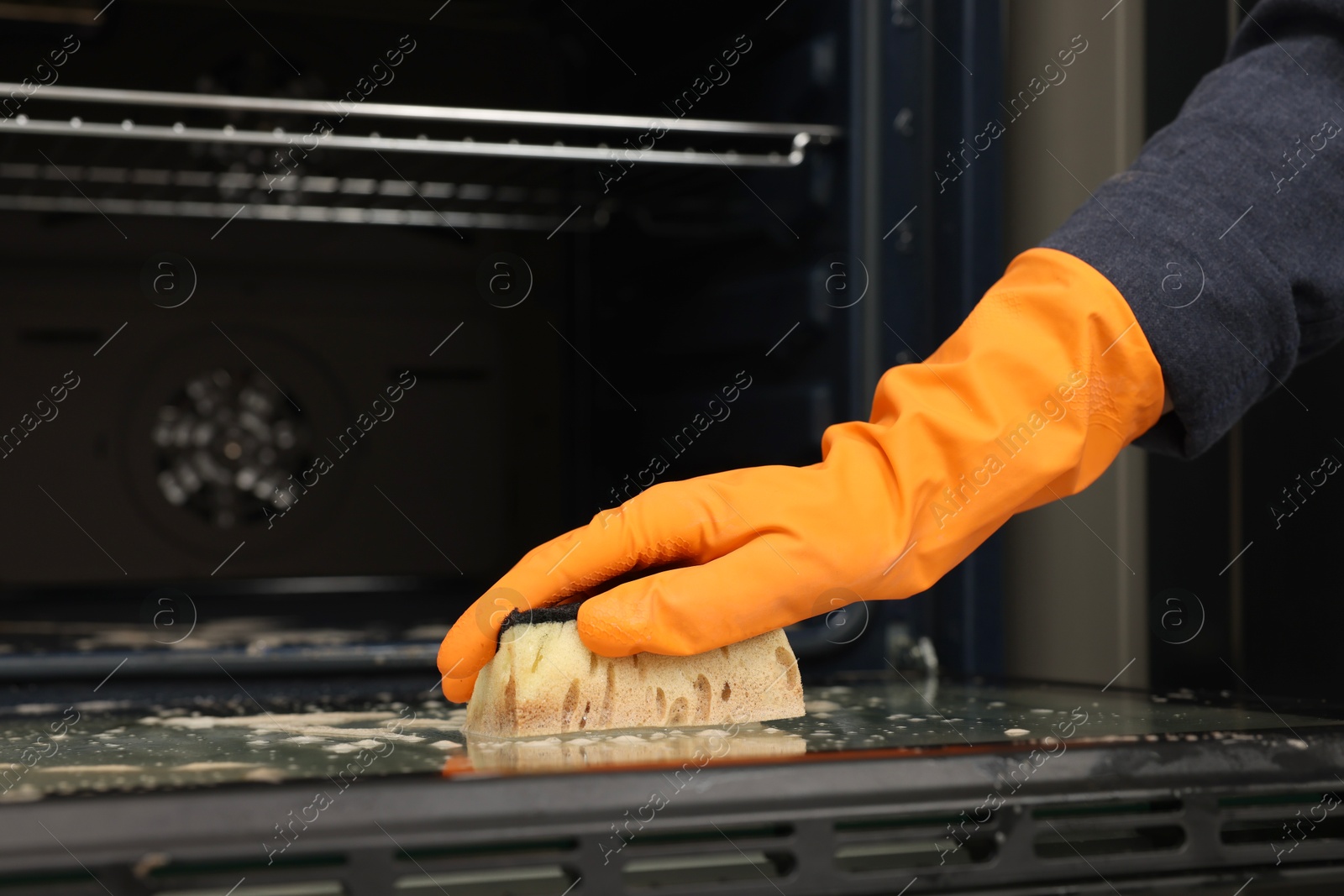 Photo of Woman cleaning oven door with sponge in kitchen, closeup