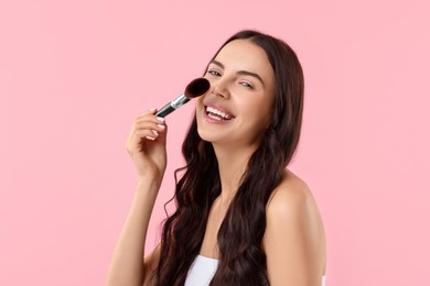 Photo of Smiling woman applying makeup with brush on pink background