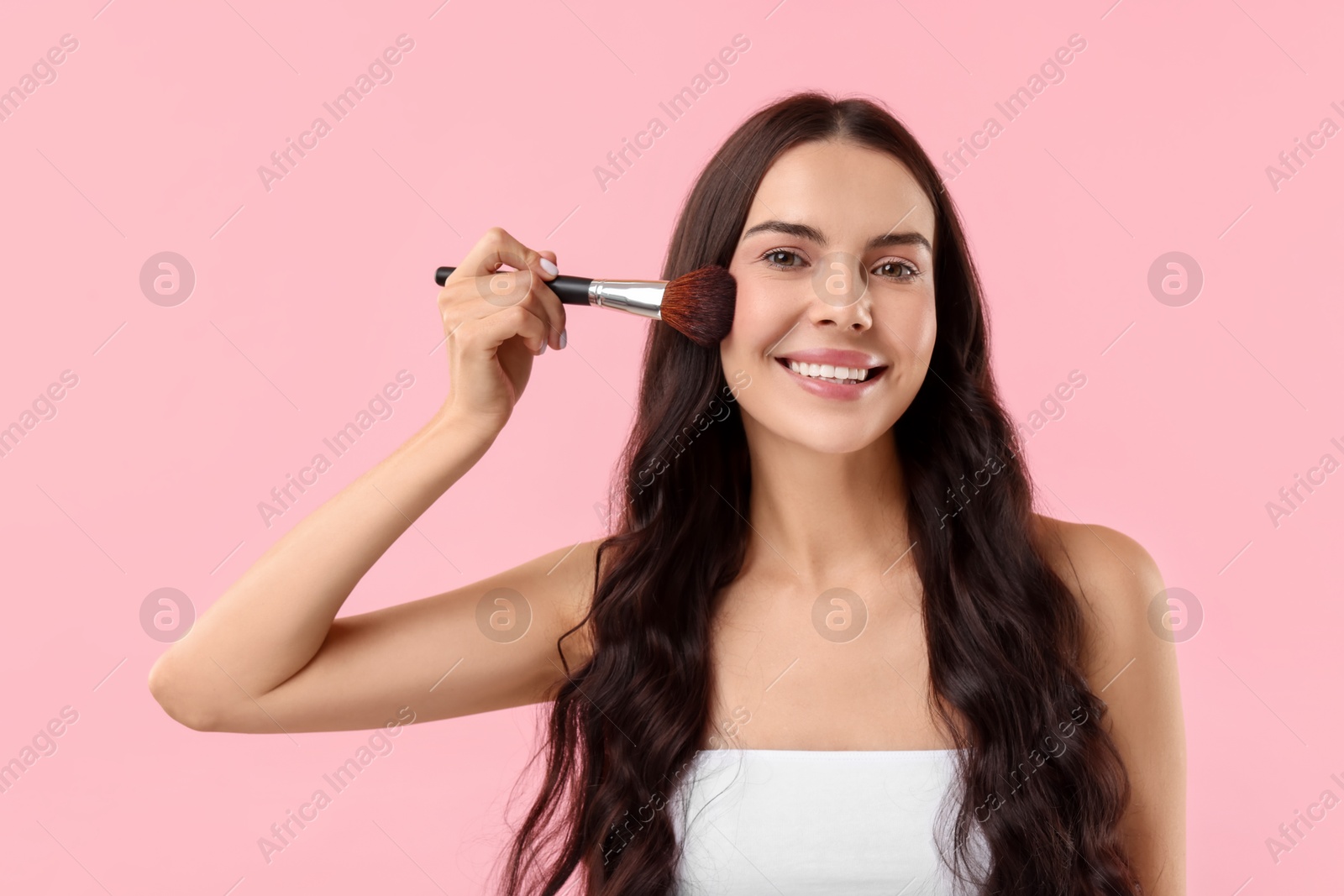 Photo of Smiling woman applying makeup with brush on pink background