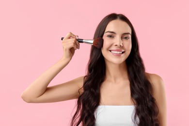 Smiling woman applying makeup with brush on pink background