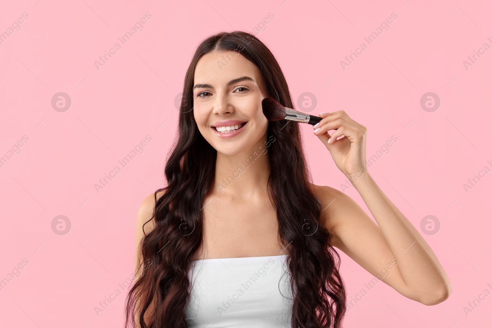 Photo of Smiling woman applying makeup with brush on pink background