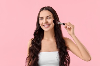 Photo of Smiling woman applying makeup with brush on pink background