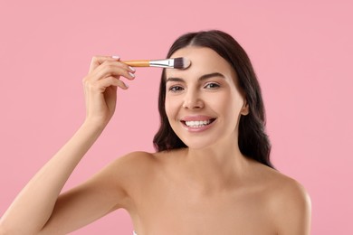 Smiling woman applying makeup with brush on pink background