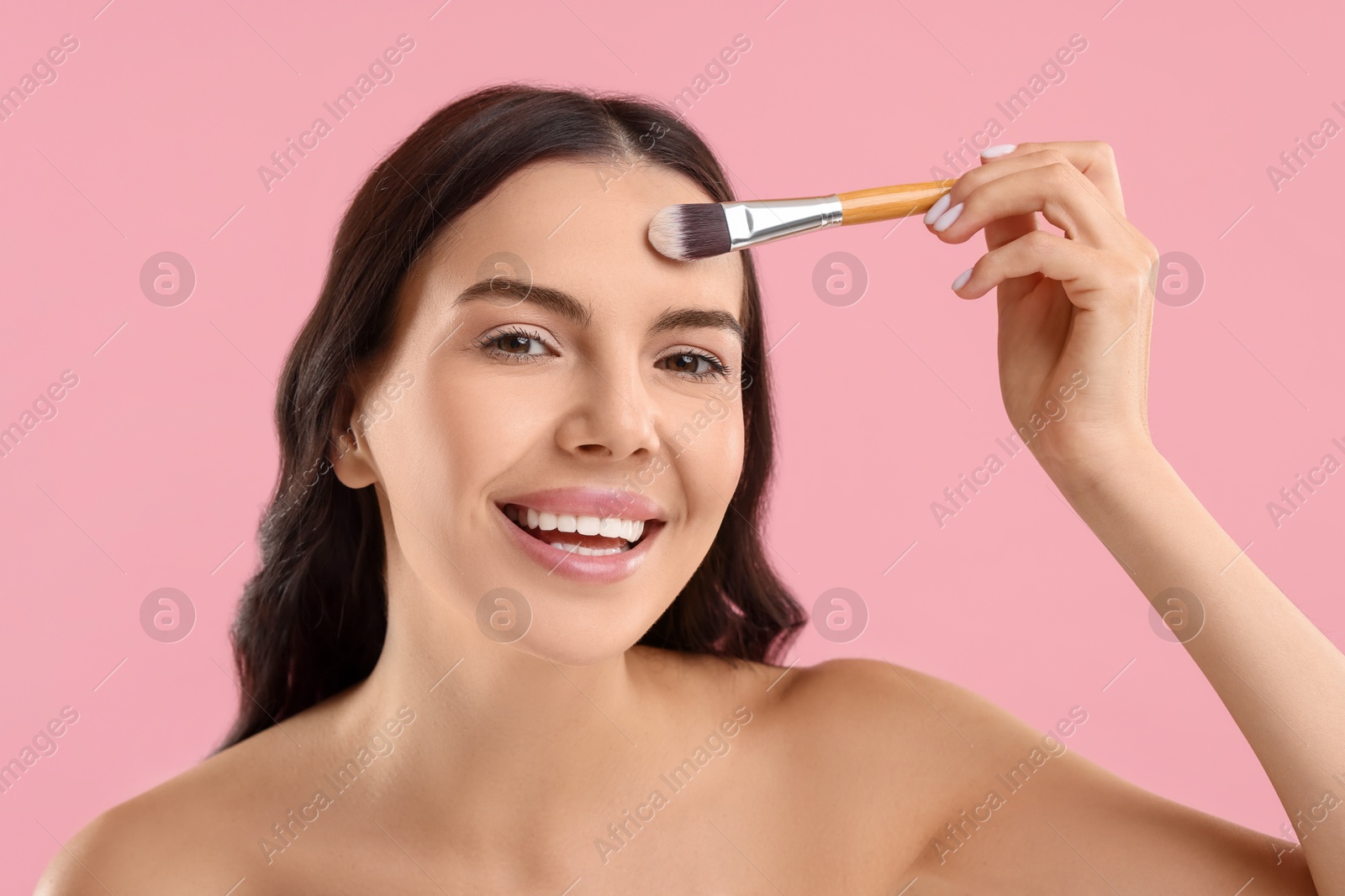 Photo of Smiling woman applying makeup with brush on pink background