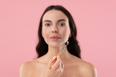 Beautiful woman applying makeup with brush on pink background