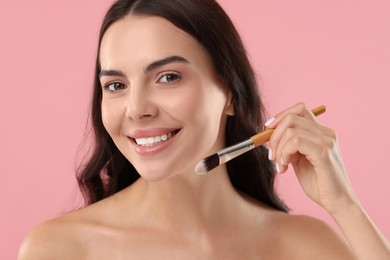 Smiling woman applying makeup with brush on pink background